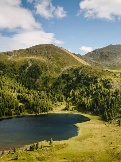 A tranquil mountain lake surrounded by green forests and gentle hills. The sky is blue and slightly cloudy. | © Erlebnisregion Murtal