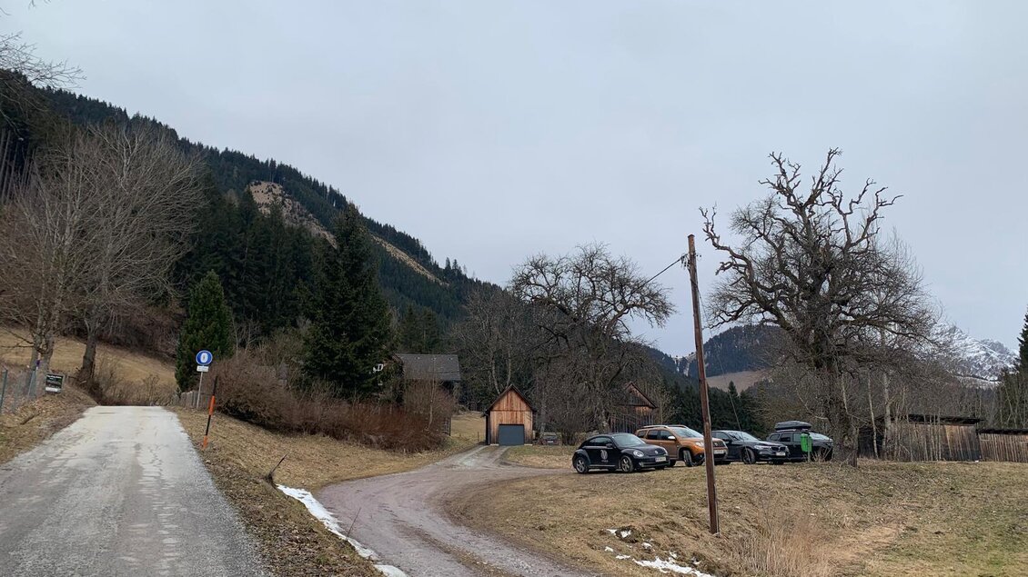 Eine ruhige ländliche Landschaft mit einem schnurgeraden Weg und einem Parkplatz. Im Hintergrund sind Bäume und Hügel sichtbar unter einem grauen Himmel. | © Barbara Pirringer