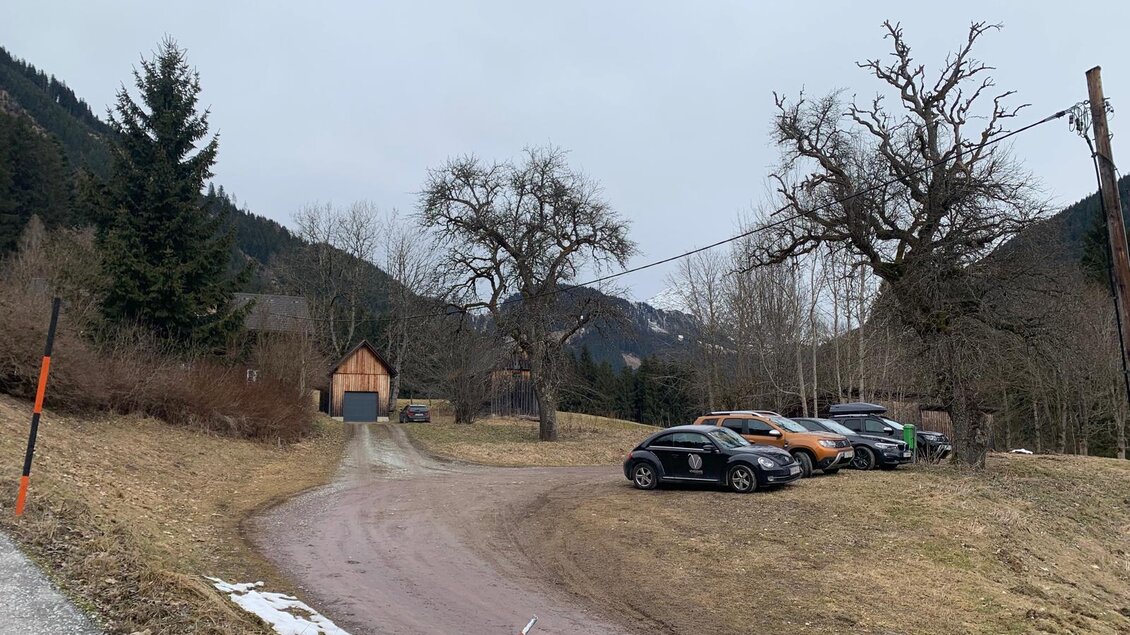 Eine ruhige Landstraße führt zu einem kleinen Haus. Mehrere Autos parken am Straßenrand, umgeben von Bäumen und sanften Hügeln. | © Barbara Pirringer