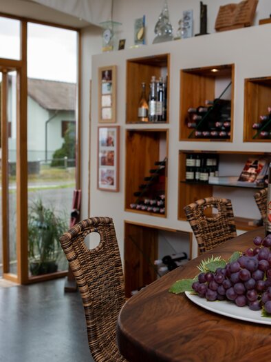 Ein einladender Weinladen mit Regalen voller Weinflaschen. Auf dem Tisch liegt ein Teller mit frischen Trauben. | © Winzerhof Klöckl