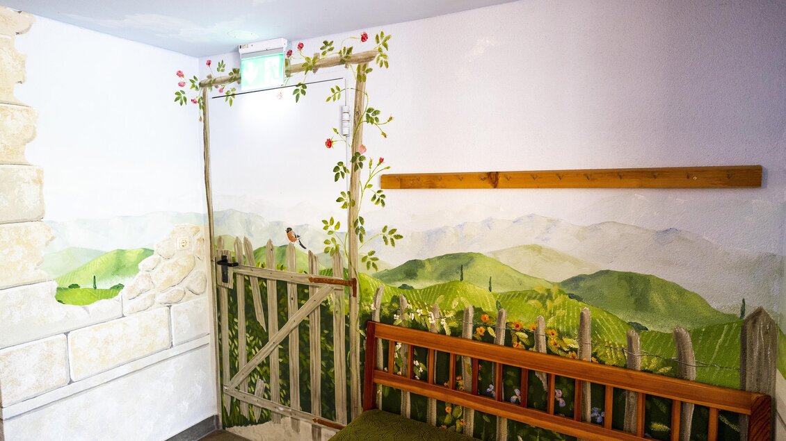 Ein gemütlicher Raum mit einer bemalten Wand, die eine ländliche Szene zeigt. Ein Holzzaun und Sanft hügelige Landschaften sind zu sehen, ergänzt durch eine Sitzbank. | © Winzerhof Strablegg