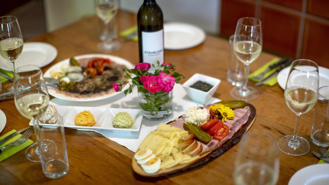 Ein schön gedeckter Tisch mit verschiedenen Gerichten, einschließlich einer Käse- und Wurstplatte. Im Hintergrund stehen Gläser mit Wein und eine Blumenvase. | © Winzerhof Strablegg