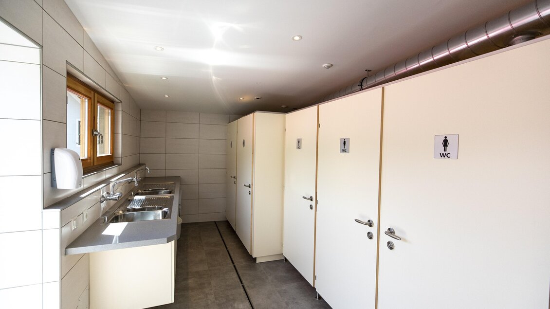 Ein helles WC mit mehreren Kabinen und Waschbecken. Die Wände sind gefliest und es gibt Fenster für Tageslicht. | © Winzerhof Strablegg