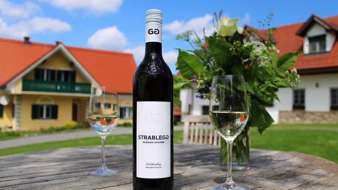 Eine Flasche weißen Weins mit der Aufschrift "STRABLEGO" steht auf einem Holztisch. Daneben stehen zwei Weingläser und im Hintergrund sind hübsche Häuser und Blumen zu sehen. | © Winzerhof Strablegg