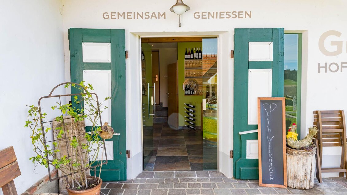 Ein einladender Eingang zu einem Weingut mit offenen Türen und einem Schild. Einige Pflanzen und Stühle auf der Veranda verleihen dem Ort eine gemütliche Atmosphäre. | © Winzerhof Strablegg