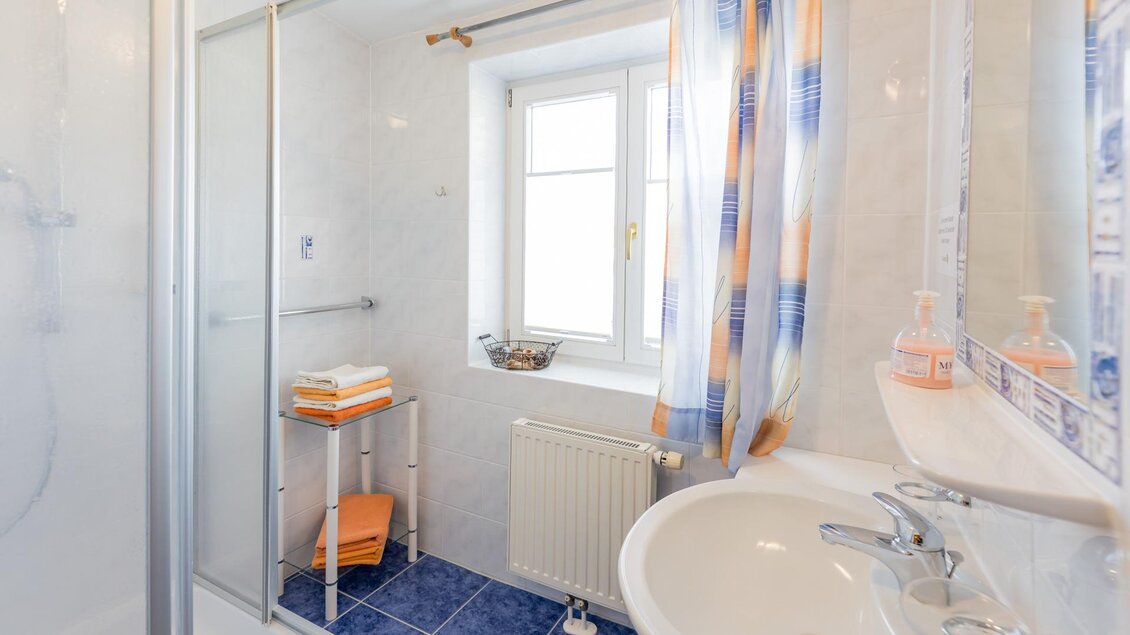 Ein modernes Badezimmer mit einer Dusche und einem Waschbecken. Das Fenster sorgt für viel Tageslicht und es gibt farbenfrohe Vorhänge. | © Winzerhof Strablegg