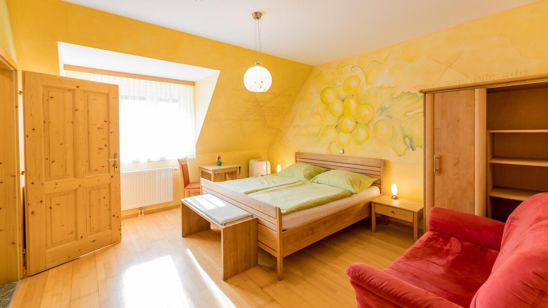 Ein gemütliches Schlafzimmer mit gelben Wänden und Holzmöbeln. Es ist hell und hat ein großes Fenster, das für natürliches Licht sorgt. | © Winzerhof Strablegg