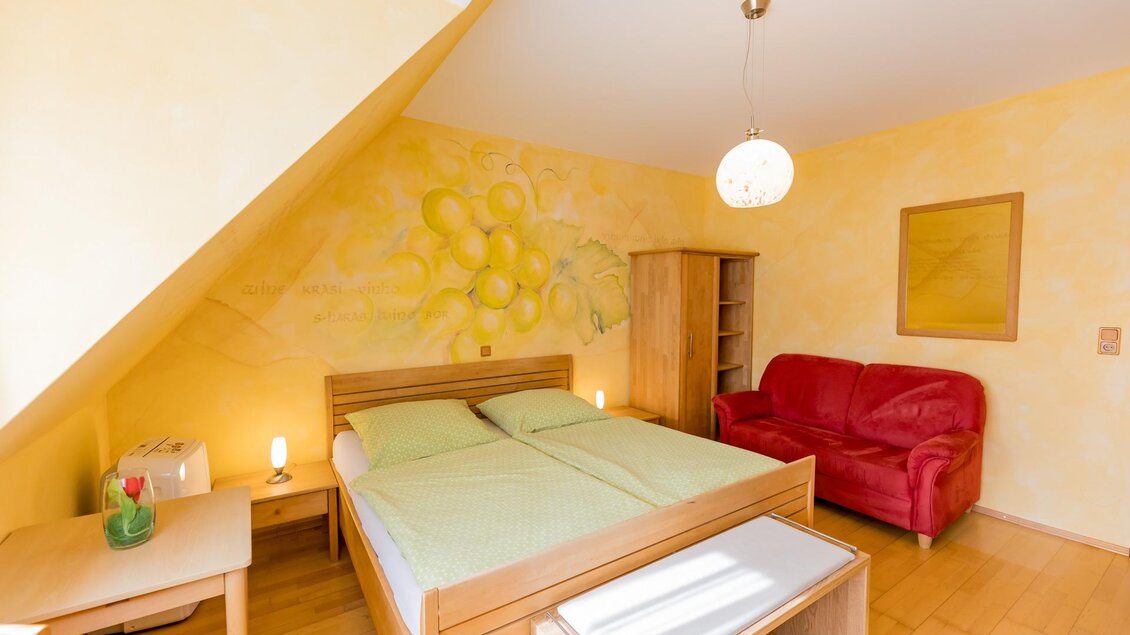 Ein gemütliches Schlafzimmer mit einem Doppelbett und einem roten Sofa. Die Wände sind in einem hellen Gelbton mit fröhlichen Dekorationen. | © Winzerhof Strablegg