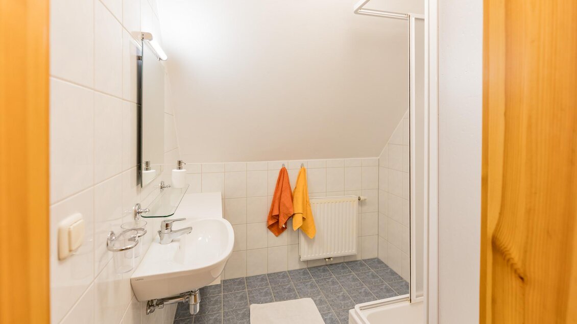 Ein modernes Badezimmer mit hellen Fliesen und einem Waschbecken. An der Wand hängen orange und gelbe Handtücher. | © Winzerhof Strablegg