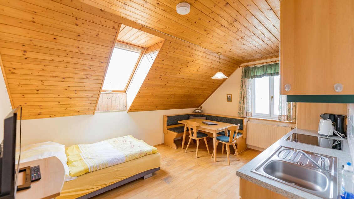 Ein gemütliches Dachzimmer mit Holzdecke und großen Fenstern. Es verfügt über ein Bett, einen Tisch und eine kleine Küchenzeile. | © Winzerhof Strablegg
