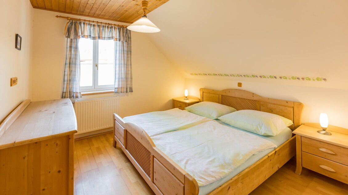Ein gemütliches Schlafzimmer mit einem großen Doppelbett und Holzmöbeln. Das Zimmer hat ein Fenster mit Vorhängen und eine warme Beleuchtung. | © Winzerhof Strablegg