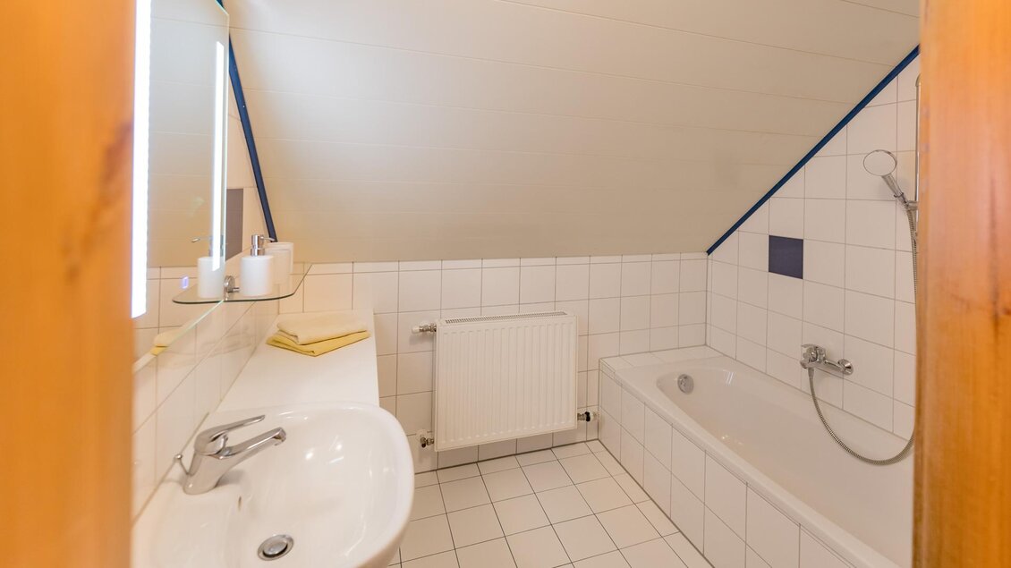 Ein helles Badezimmer mit weißer Fliesenwand und einem Waschbecken. Auf der rechten Seite befindet sich eine Badewanne. | © Winzerhof Strablegg