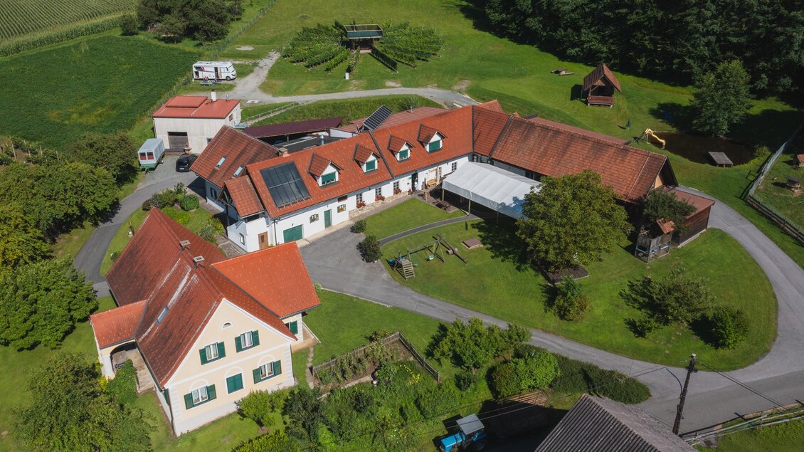 Eine weitläufige Ansicht eines Bauernhofs mit mehreren Gebäuden und umgebenden Wiesen. Der Hof ist von grünen Bäumen und Sträuchern umgeben. | © Winzerhof Strablegg