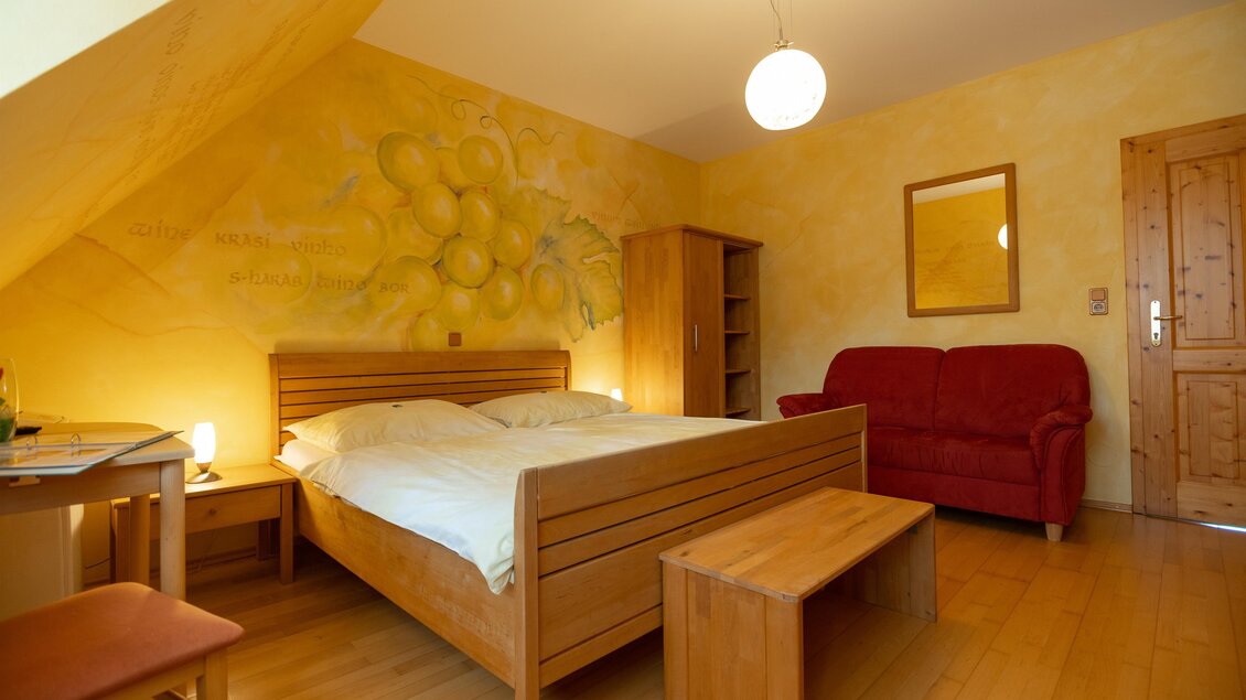 Ein gemütliches Schlafzimmer mit einem großen Bett und einer Sofaecke. Die Wände sind hellgelb gestrichen und mit einer Obstillustration dekoriert. | © Winzerhof Strablegg