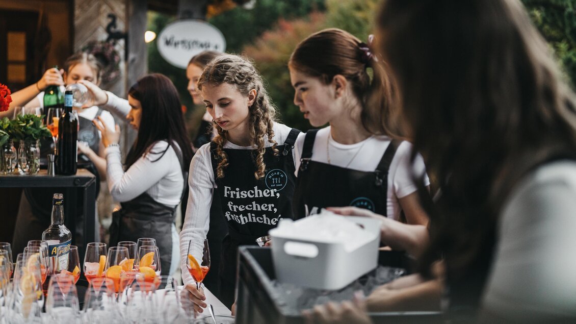 Eine Gruppe junger Frauen bereitet Getränke an einem Bartresen vor. Im Hintergrund sind gläserne Cocktails und frische Zutaten zu sehen. | © Heldentheater