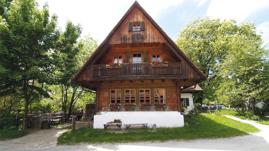 Ein traditionelles Holzhaus mit einem spitzen Dach und vielen Fenstern. Es steht in einer grünen Umgebung mit Bäumen und einem kleinen Garten. | © Jagawirt
