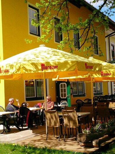 A cozy café with sunny terrace seats under a yellow sun umbrella.  
Guests enjoy the fresh air in a green environment. | © Anbauer