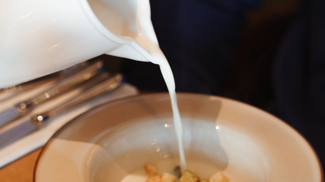 Eine Person gießt Milch in eine Schüssel mit kleinen Würfeln und einem grünen Blatt. Der Hintergrund zeigt eine entspannte Essensatmosphäre. | © Yvonne Bernhard
