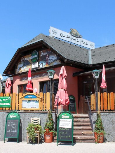 A cozy restaurant with an inviting facade. Red and white umbrellas stand in front of the entrance along with signs displaying the menu offerings. | © TVB Ausseerland - Salzkammergut_Viola Lechner