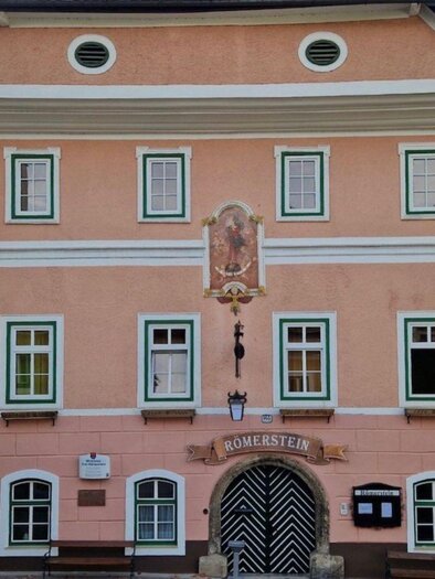 <p>An inviting inn with a historic facade displaying the name "Römerstein" and a distinctive two-colored door. The pink facade is cheerful and features green window frames.</p> | © Gerhard Fallent