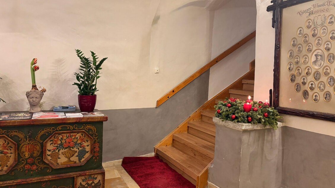 Ein großzügiger Eingangsbereich mit hellen verputzten Wänden, einem schön bemalten Holzschrank, und liebevoller Dekoration wie ein Pflanztrog und Blumen. Eine breite Holztreppe führt in das Obergeschoss, davor befindet sich ein edler roter Teppich. | © Wirtshaus zum Römerstein