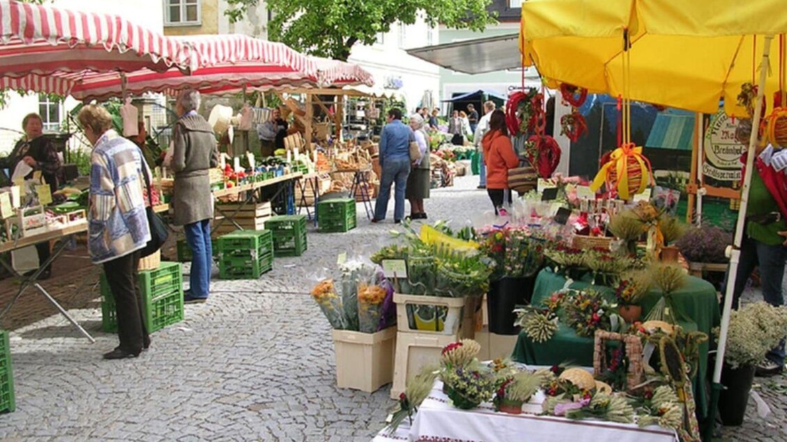 Der lebhafte Ausseer Wochenmarkt mit bunten Ständen und frischen Lebensmitteln sowie Blumen im Sommer. Besucher stöbern zwischen verschiedenen Ständen und kaufen lokale Produkte. | © Stadtgemeinde Bad Aussee