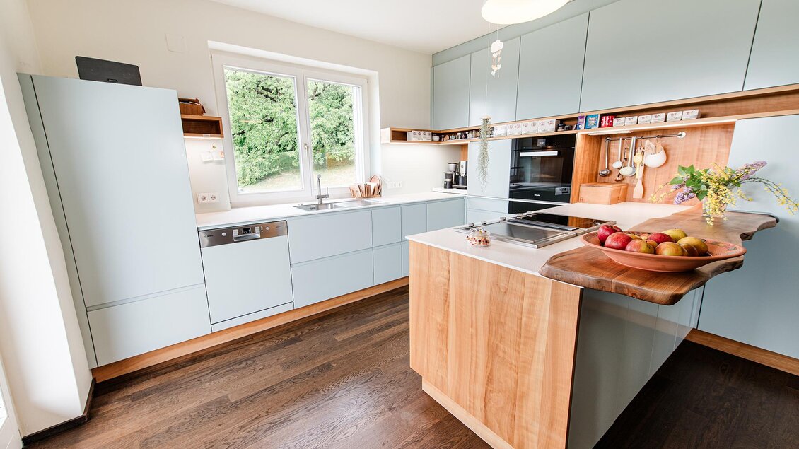 Eine moderne Küche mit hellblauen Schränken und Holzdetails. Auf der Kücheninsel liegen frisches Obst und Blumen. | © (c) Steirisches Vulkanland / Manuel Flor