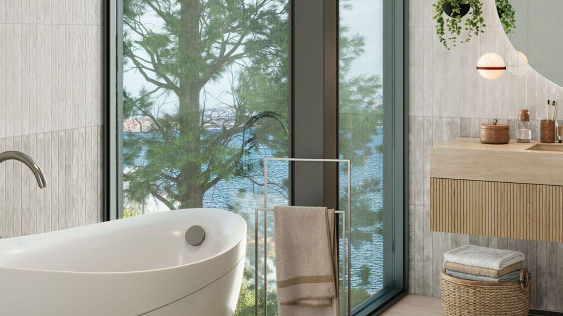 Ein modernes Badezimmer mit einer freistehenden Badewanne und großen Fenstern, die einen Blick auf Bäume und Wasser bieten. Der Raum ist hell und minimalistisch eingerichtet. | © Archiv Wohnkeramik-Lukas Tanzmeister