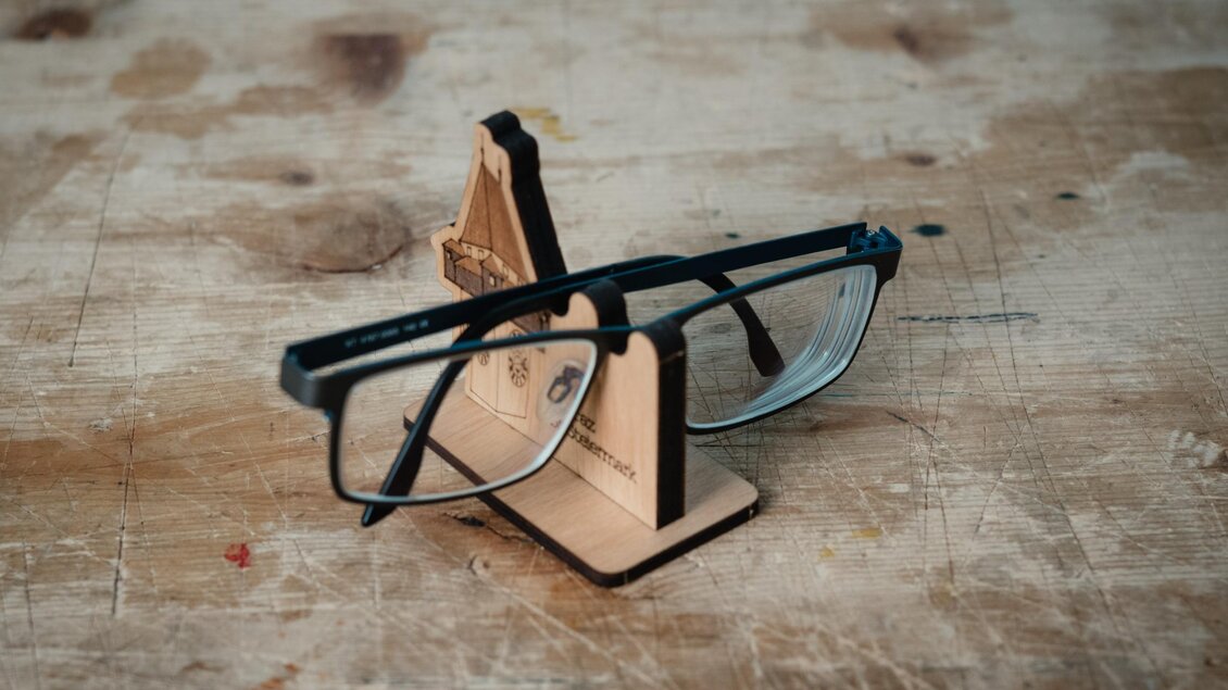 Schwarze Brille liegt auf einem gravierten Brillenhalter aus Holz von WoodHeroes in Graz. | © Bernd Grosseck