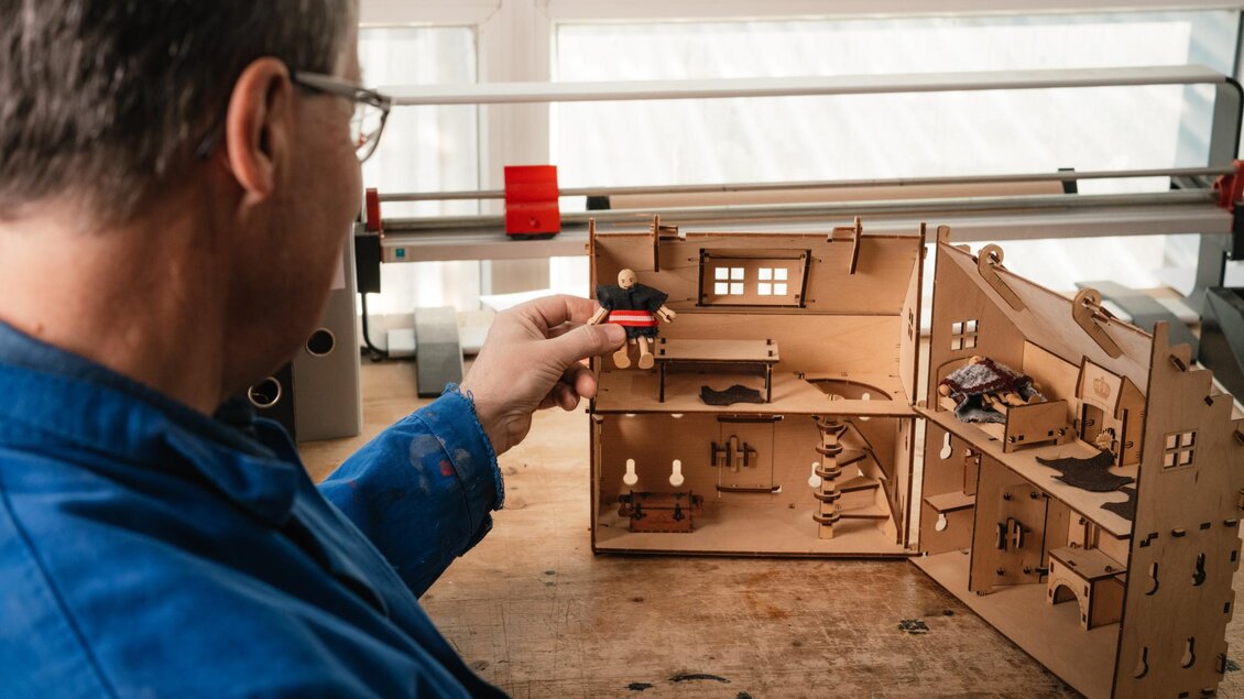 Ein Mann hält eine Spielfigur vor ein geöffnetes Spielhaus aus Holz mit mehreren Räumen und kleinen Möbeln. | © Bernd Grosseck