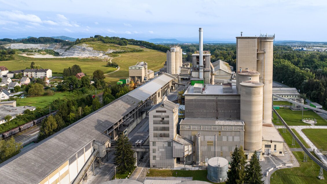 Eine große Industrieanlage mit mehreren Fabrikationsgebäuden und Schornsteinen. Im Hintergrund sind Hügel und eine grüne Landschaft zu sehen. | © Robert Sommerauer