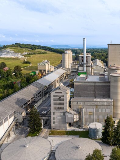 Eine große Industrieanlage mit mehreren Fabrikationsgebäuden und Schornsteinen. Im Hintergrund sind Hügel und eine grüne Landschaft zu sehen. | © Robert Sommerauer