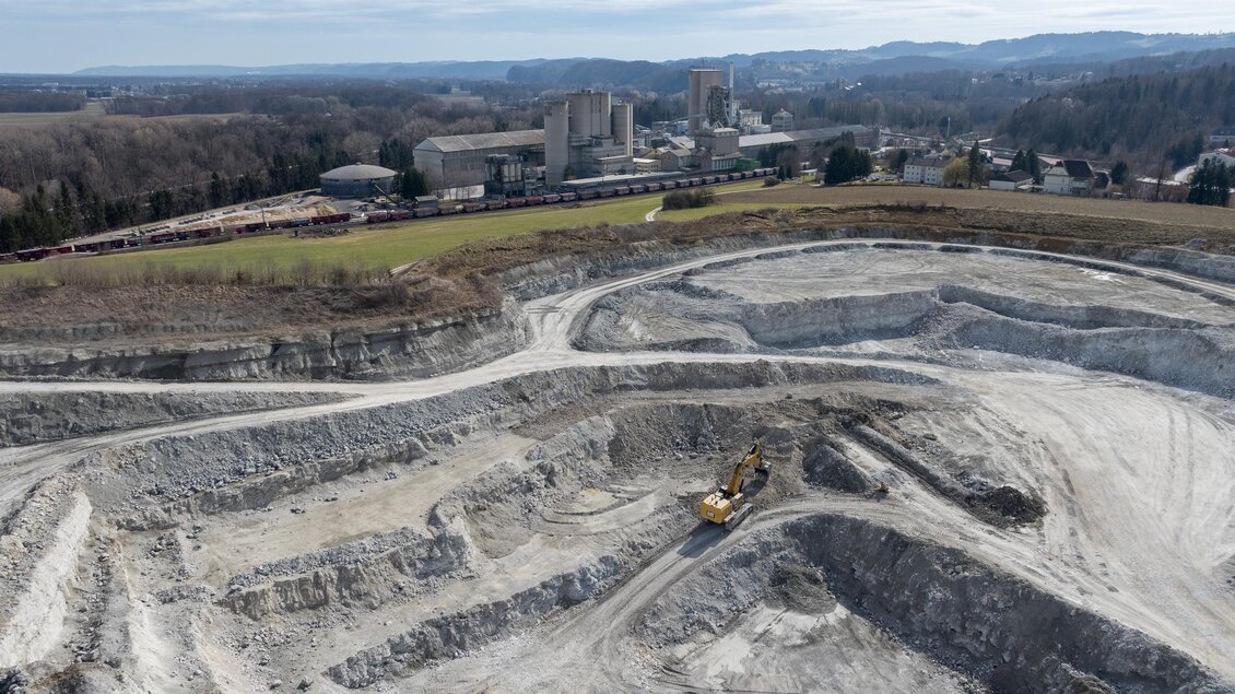 Eine große Baustelle mit verschiedenen Erdhügeln und einem Bagger. Im Hintergrund sind Fabrikgebäude und eine hügelige Landschaft zu sehen. | © Robert Sommerauer