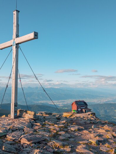 Ein Gipfel mit einem großen Kreuz und einer kleinen Hütte. Im Hintergrund erstrecken sich die Berge und der Himmel ist klar und blau. | © TVB Murau
