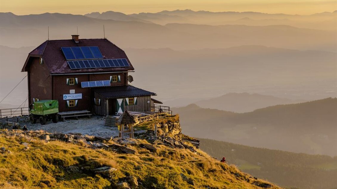 Eine Berghütte mit Solarpanelen auf einem Hügel. Im Hintergrund sind sanfte Berge und ein farbenfroher Sonnenuntergang zu sehen. | © Erlebnisregion Murtal