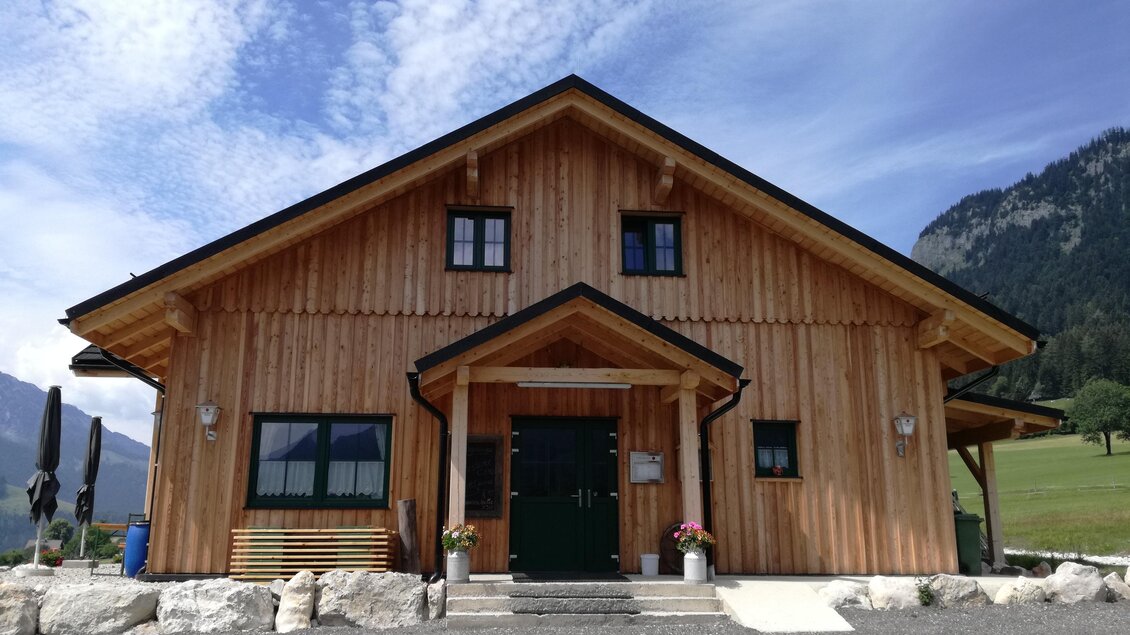 <p>Das schöne Holzhaus von „Zum fliegenden Honigtopf“ mit einer großen Eingangstür und Fenstern. Im Hintergrund sind grüne Wiesen und die Berge des Ausseerlandes zu sehen.</p> | © Stoffis Hütten
