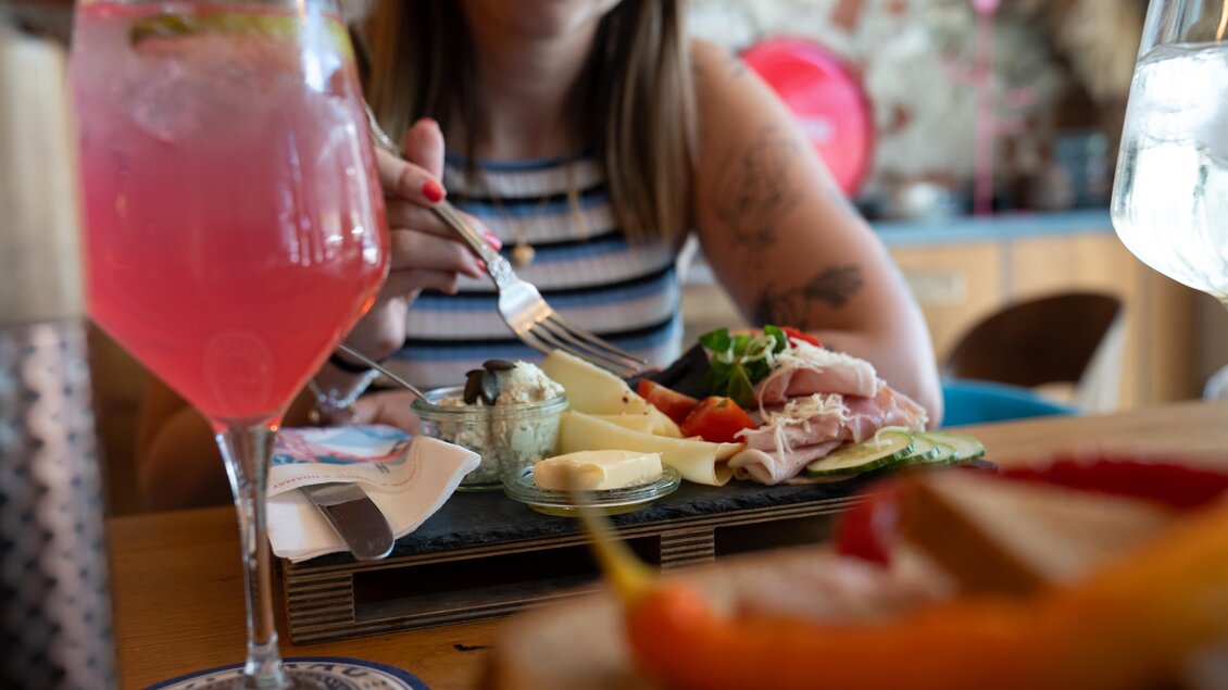 Eine Person isst eine bunte Platte mit frischem Gemüse, Aufschnitt und Käse. Im Vordergrund stehen zwei Getränke, ein pinkes Getränk und ein Glas Wasser. | © STG