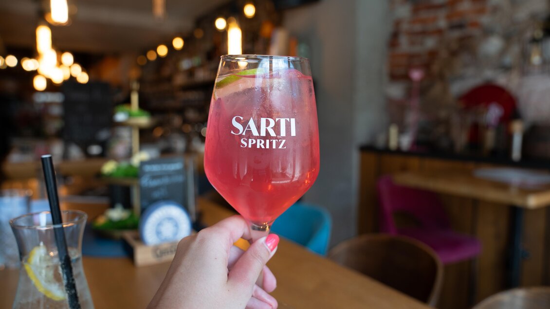 Ein frischer Sarti Spritz in einem eleganten Glas, gehalten von einer Hand. Im Hintergrund sind stilvolle Möbel und eine gemütliche Atmosphäre zu sehen. | © STG