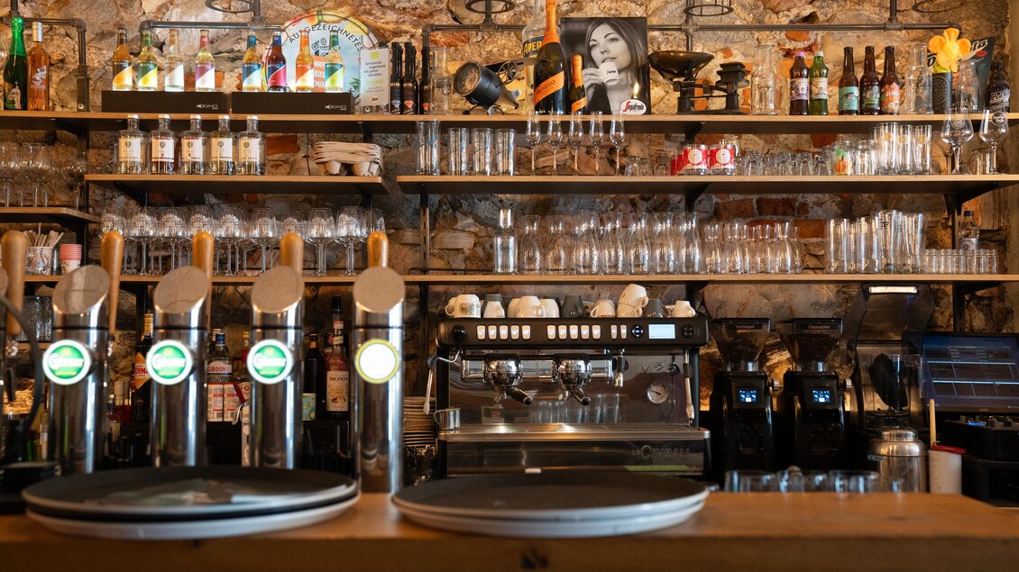 Eine gemütliche Bar mit Holzregalen und einer vielfältigen Getränkeauswahl. Im Hintergrund sind Gläser und eine Kaffeemaschine zu sehen. | © STG