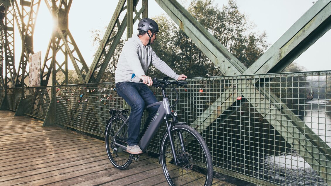 Ein Radfahrer steht auf einer Holzbrücke und schaut auf die Landschaft. Die Brücke hat eine grüne Metallstruktur und im Hintergrund sind Bäume sichtbar. | © RM SWS GmbH | webquartier.at