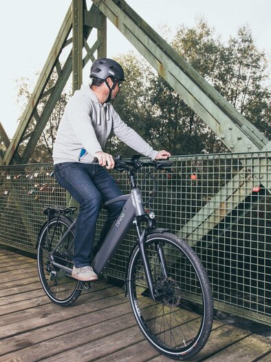 Ein Radfahrer steht auf einer Holzbrücke und schaut auf die Landschaft. Die Brücke hat eine grüne Metallstruktur und im Hintergrund sind Bäume sichtbar. | © RM SWS GmbH | webquartier.at