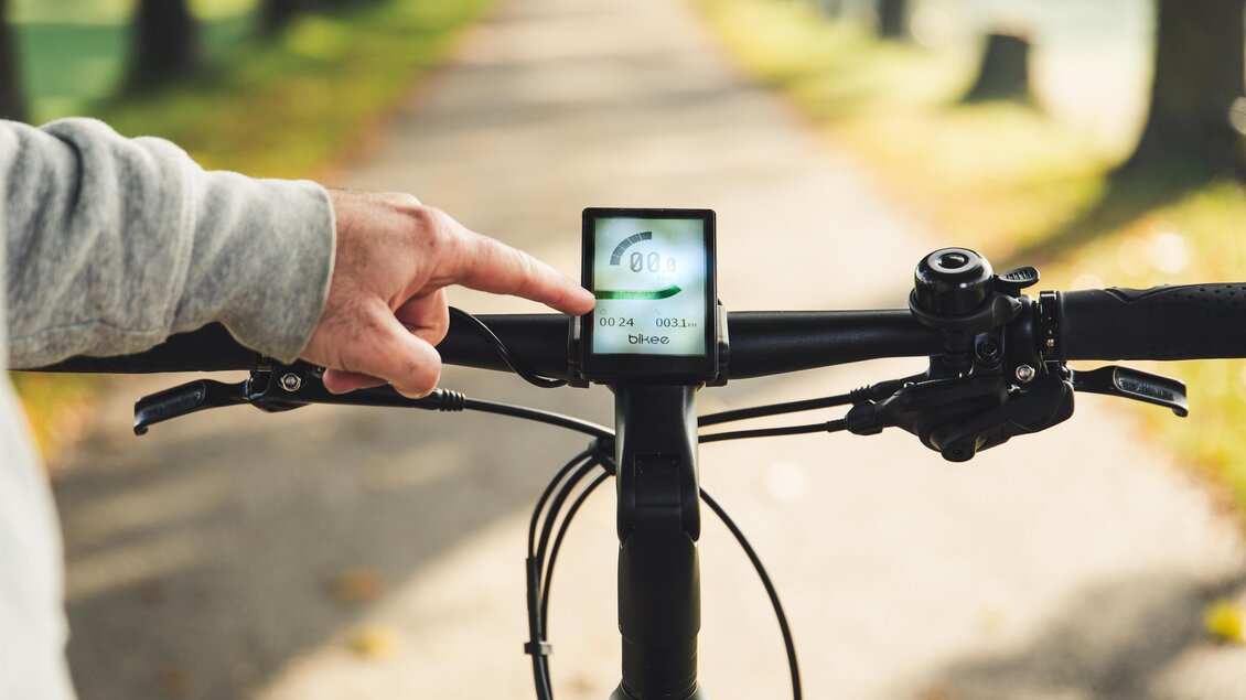 Ein Fahrradlenker mit einem Display, auf dem Einstellungen angezeigt werden. Eine Hand tippt auf das Display, während der Weg im Hintergrund sichtbar ist. | © RM SWS GmbH | webquartier.at