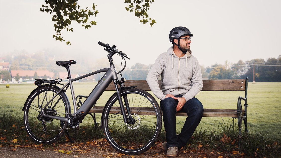 Ein Mann sitzt auf einer Bank neben seinem E-Bike. Die Umgebung ist ruhig und ländlich mit Bäumen im Hintergrund. | © RM SWS GmbH | webquartier.at
