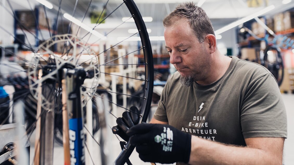 Ein Mann arbeitet konzentriert an einem Fahrradreifen in einer Werkstatt. Im Hintergrund sind zahlreiche Fahrräder und Ausrüstungen zu sehen. | © RM SWS GmbH | webquartier.at