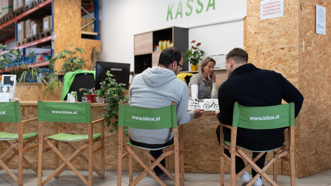 Ein Verkaufsbereich mit einem Empfangstresen und Pflanzen. Zwei Personen sitzen auf grünen Stühlen und schauen auf ein Menü. | © RM SWS GmbH | webquartier.at