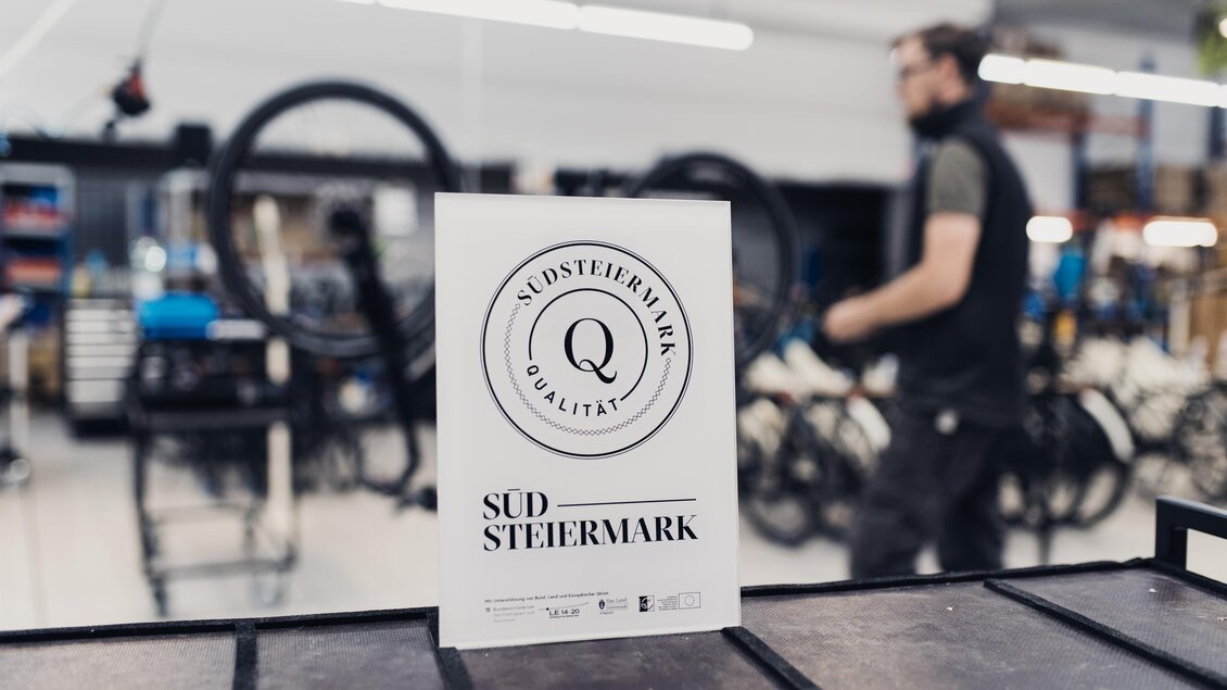 Ein Schild mit dem Text „Südsteiermark Qualität“ steht auf einem Tisch in einer Werkstatt. Im Hintergrund sind Fahrräder und eine Person, die beschäftigt ist, zu sehen. | © RM SWS GmbH | webquartier.at
