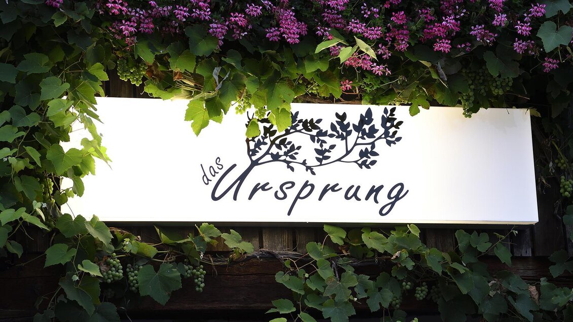 Ein Schild mit der Aufschrift "das Ursprung" ist umgeben von grünem Efeu und pinken Blumen. Die Begrünung verleiht dem Schild einen einladenden und natürlichen Look. | © Das Ursprung