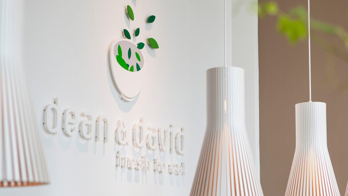 Ein modernes Restaurantlogo von "dean & david" an einer hellen Wand. Im Hintergrund hängen elegante, weiße Lampen. | © dean&david