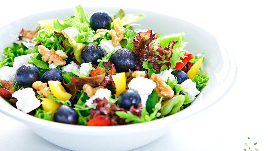 Ein frischer Salat mit verschiedenen grünen Blattsalaten, Heidelbeeren, Nüssen und buntem Gemüse. Obenauf sind weiße Käsewürfel zu sehen. | © dean&david
