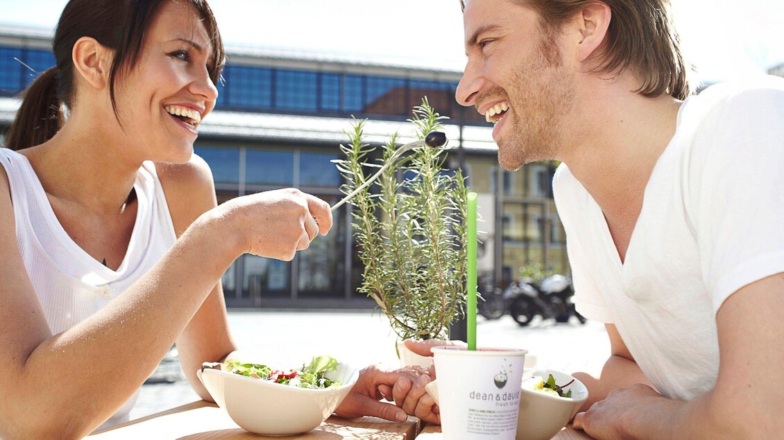 Ein fröhliches Paar genießt ein gesundes Essen im Freien. Sie lachen und teilen sich eine Salatschüssel. | © dean&david